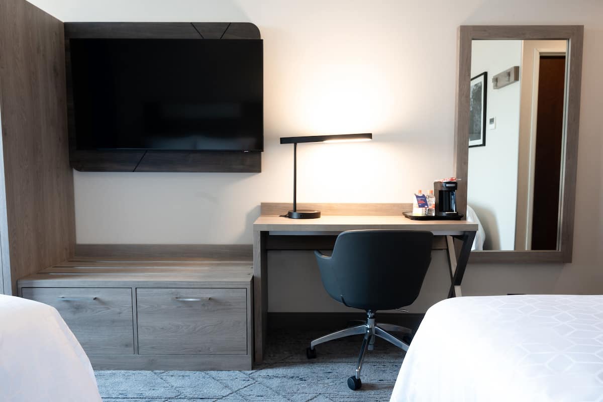 A hotel room with a bed, a desk with a chair, a flat-screen TV mounted on the wall, a desk lamp, and a coffee maker.
