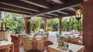 An outdoor dining area with tables set for guests, surrounded by lush greenery and a pool. - Hotel San Marco (Santo Domingo)