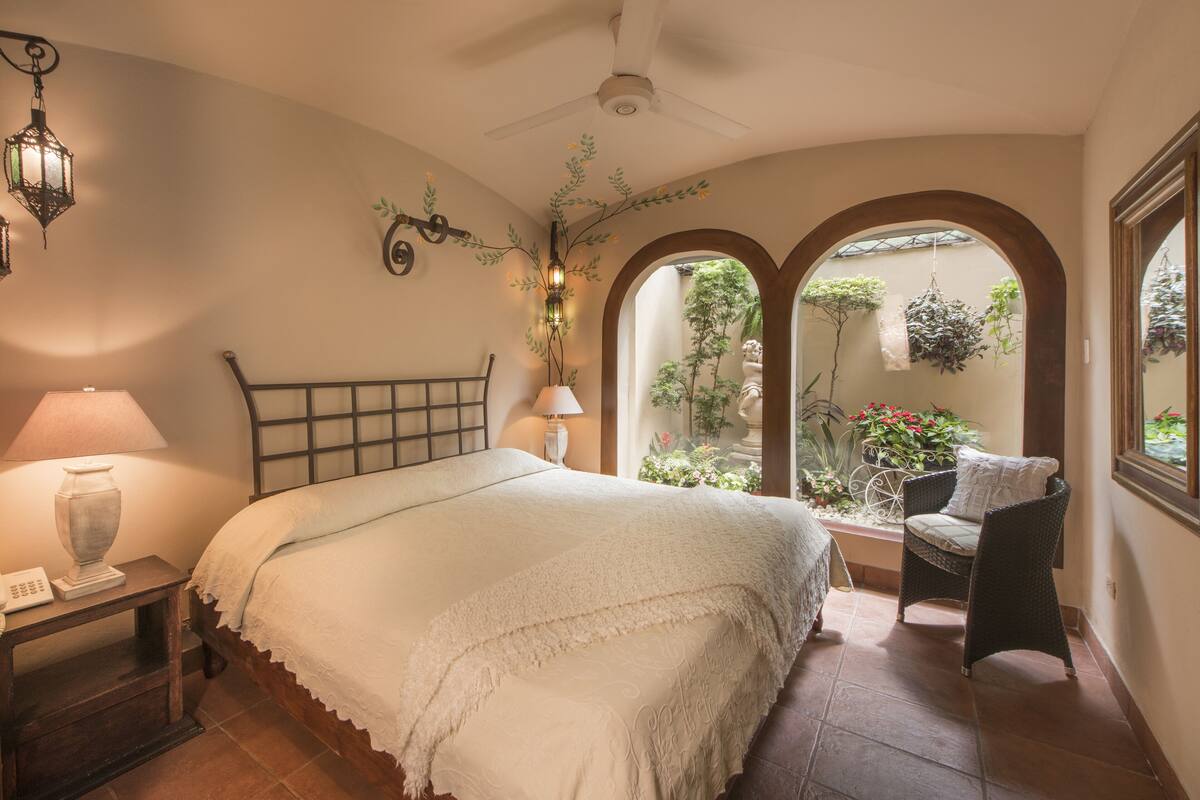 A bedroom with a bed, bedside table, lamp, and a view of a garden through an arched window.