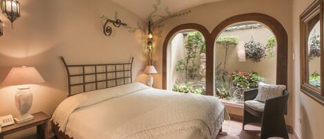 A bedroom with a bed, bedside table, lamp, and a view of a garden through an arched window.