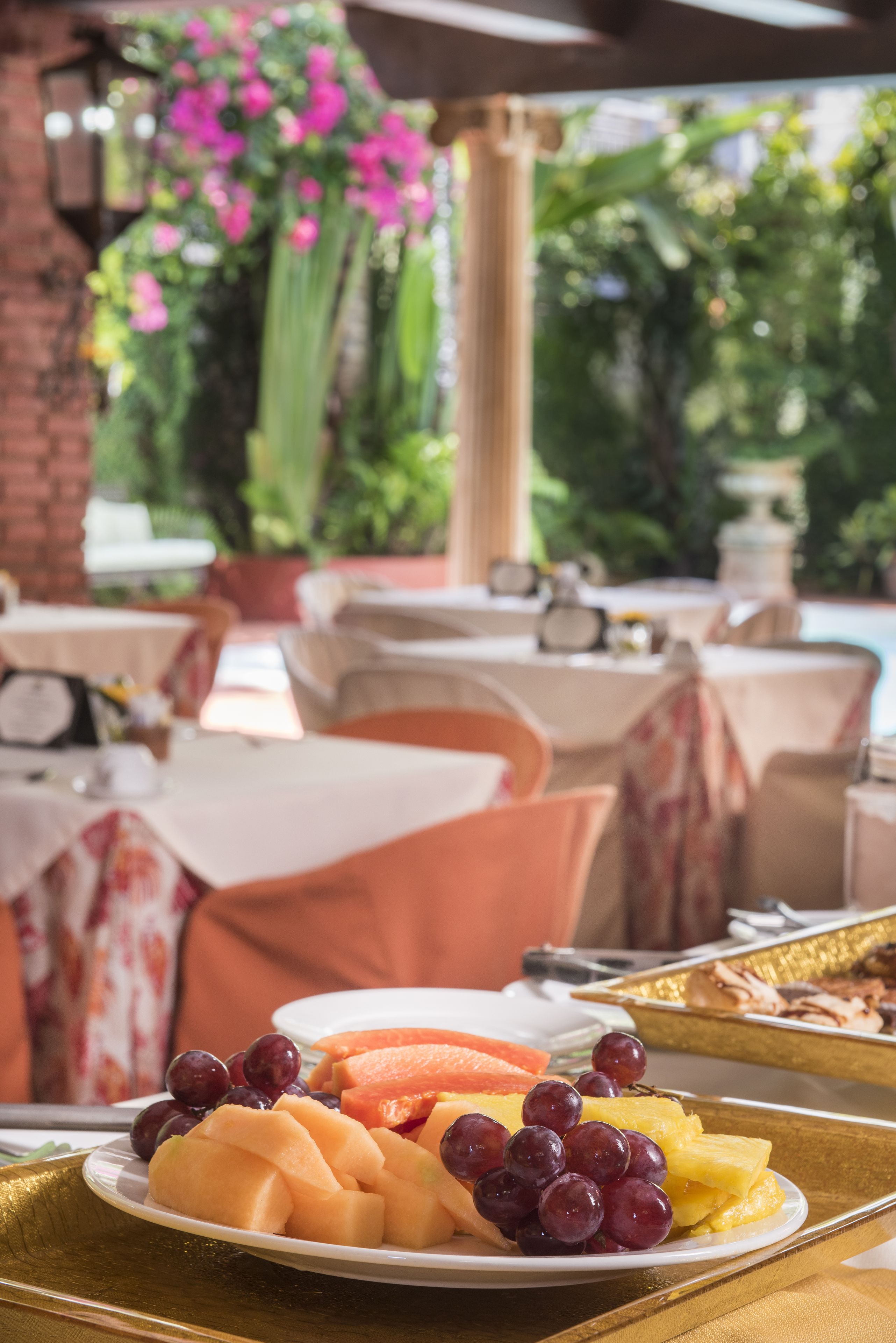 A table with a plate of fruit and a tray of food, set in an outdoor dining area with tables and chairs.
