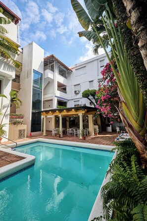 A pool area with a covered seating area, surrounded by lush greenery and a multi-story building with balconies.