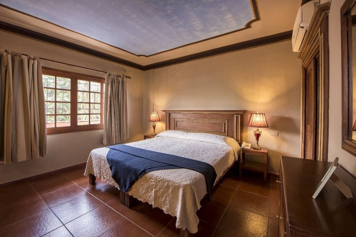A bedroom with a large bed, two bedside lamps, a window with curtains, and a ceiling skylight.