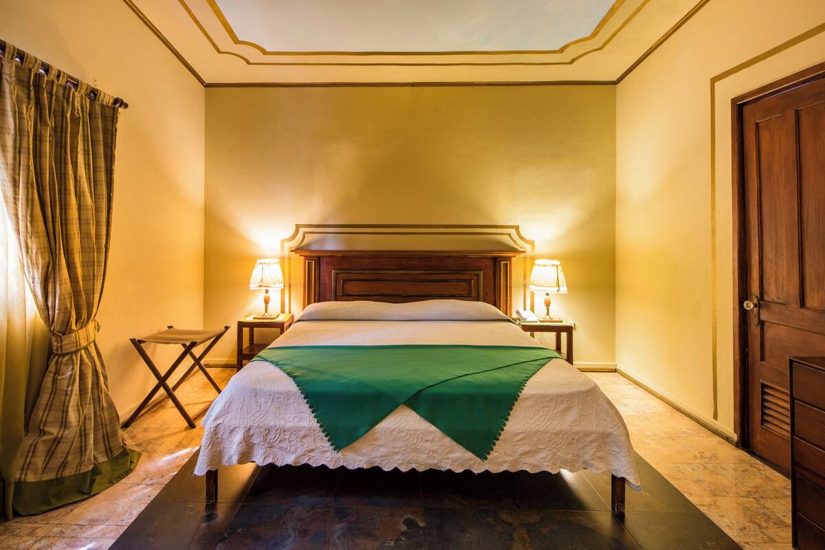 A neatly made bed with a green throw, two bedside lamps, and a wooden headboard in a room with yellow walls and a wooden door.