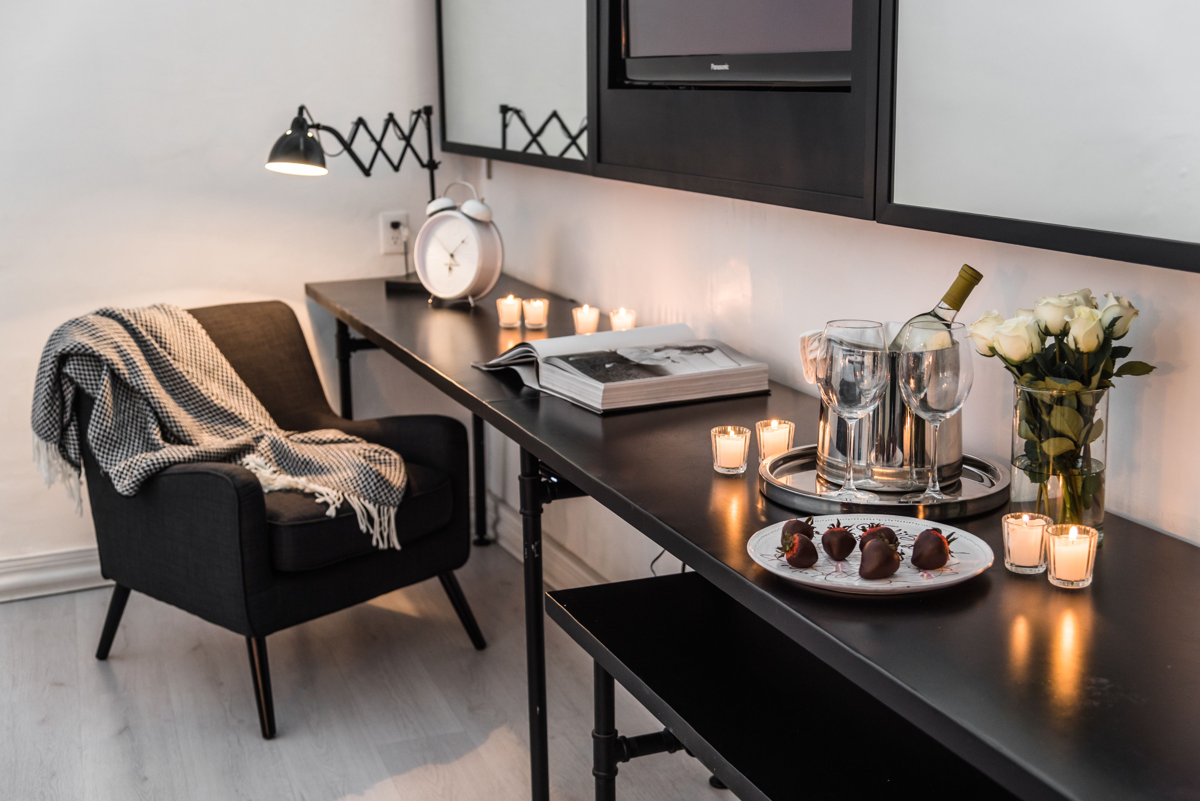 A modern room with a black sofa, a small table with candles, a clock, a tray with chocolates, and a vase with flowers.