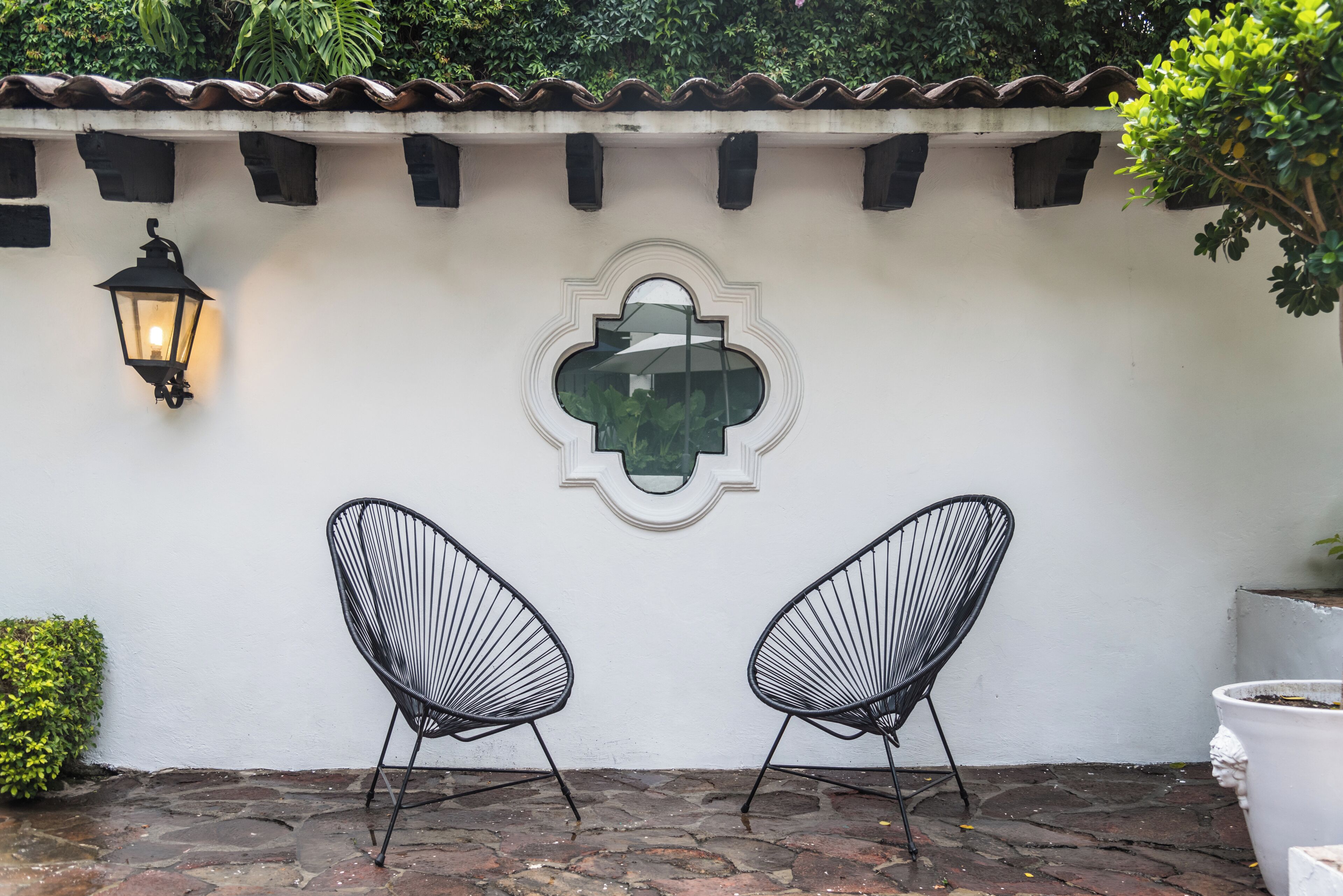 Dos sillas de alambre negro frente a una pared blanca con un ventanal decorativo y un farol de pared.