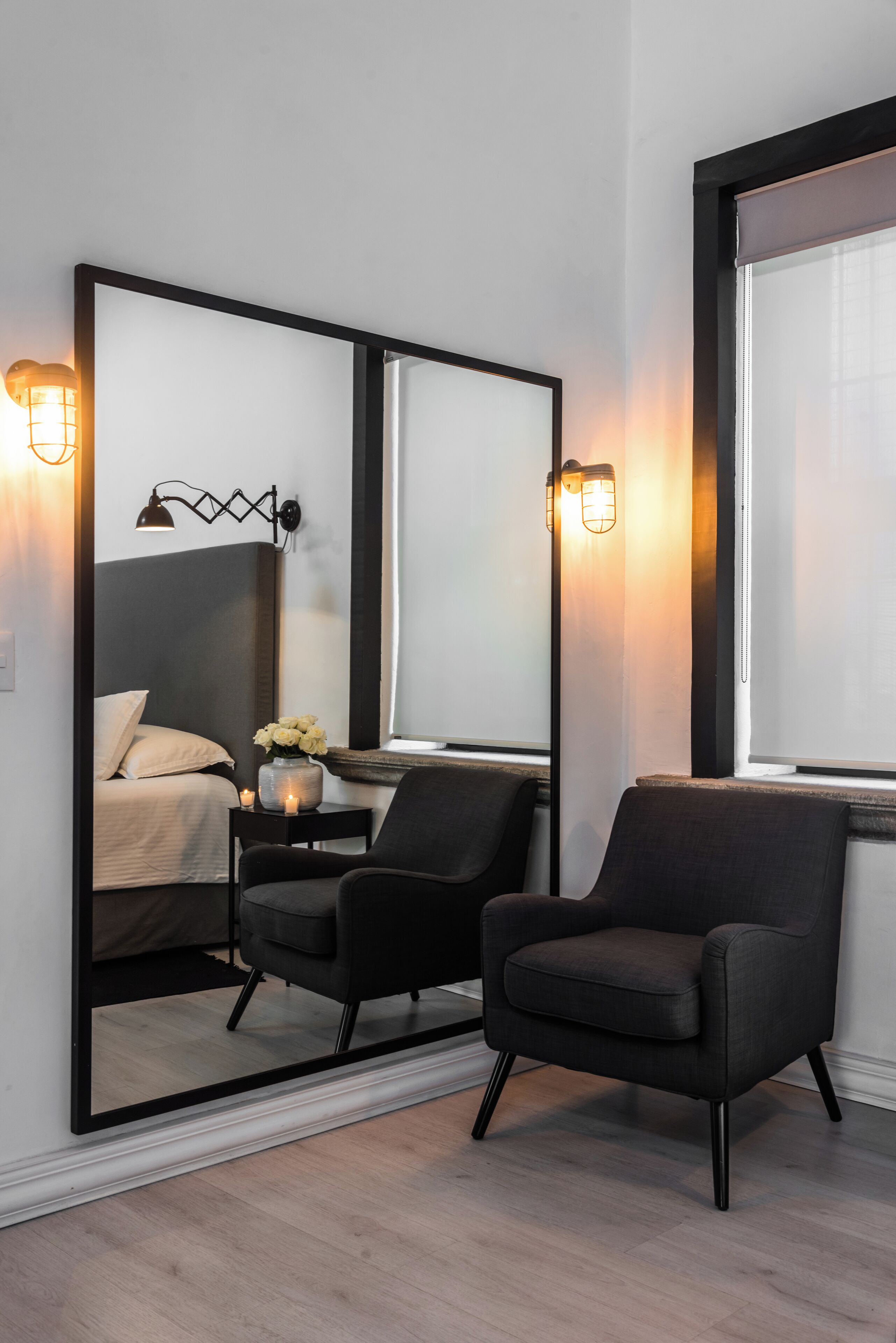 Una habitación de hotel moderna con un espejo grande, dos sillones negros, una mesita de noche con un jarrón de flores y una cama con ropa blanca.