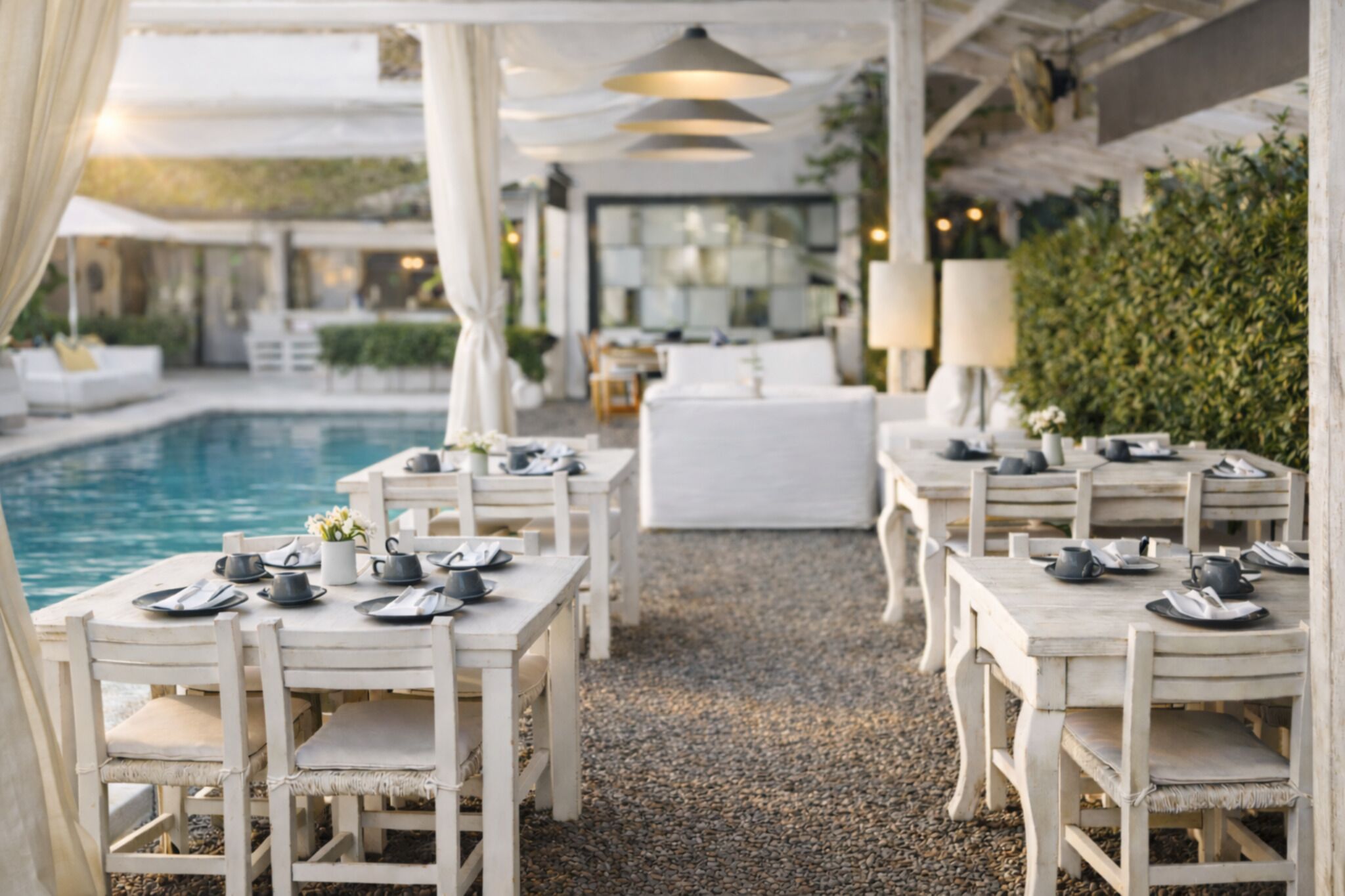 Un área de comedor al aire libre con mesas y sillas blancas, una piscina y un seto.