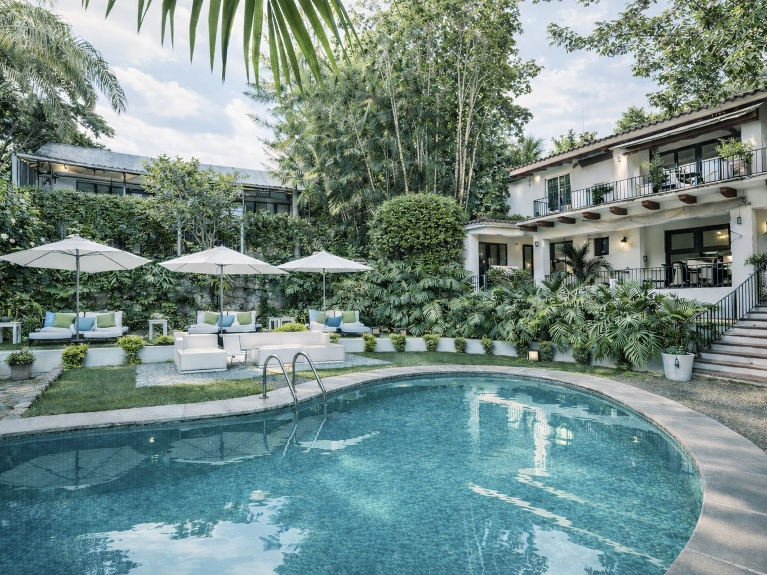 A pool area with lounge chairs and umbrellas, surrounded by lush greenery and a two-story building with balconies.