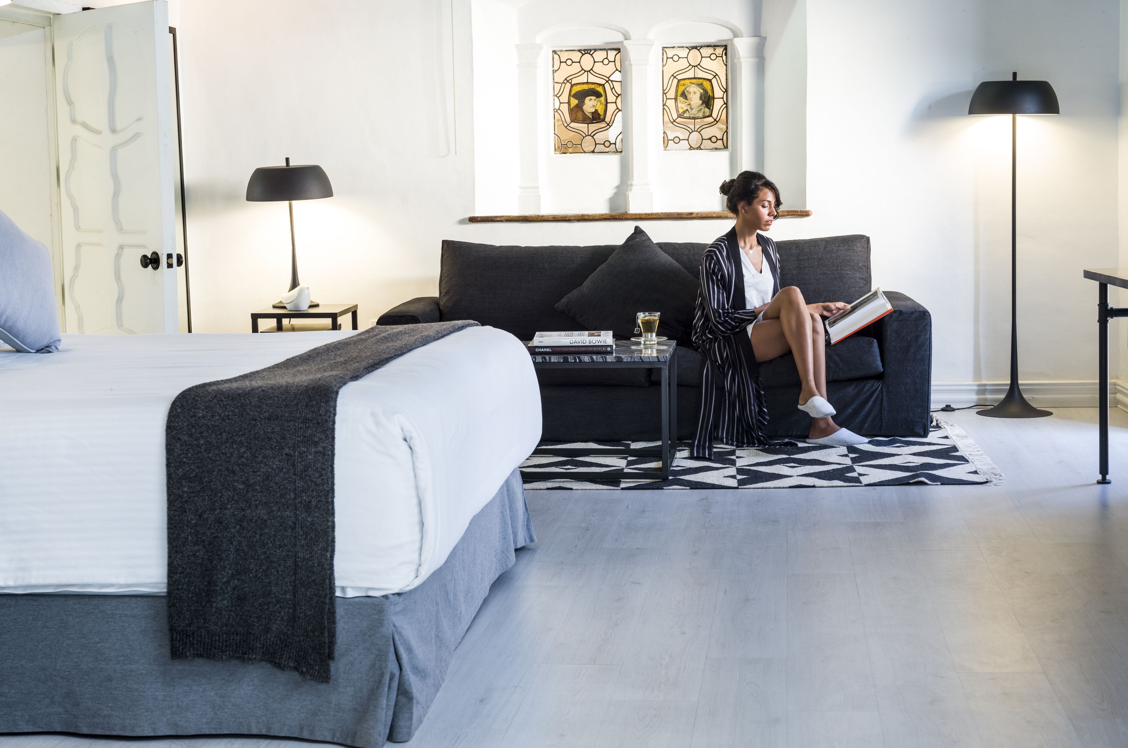 Una habitación de hotel moderna con una cama grande, un sofá y una mesita con libros y una copa de vino. Hay dos marcos de cuadros en la pared y una lámpara de pie.