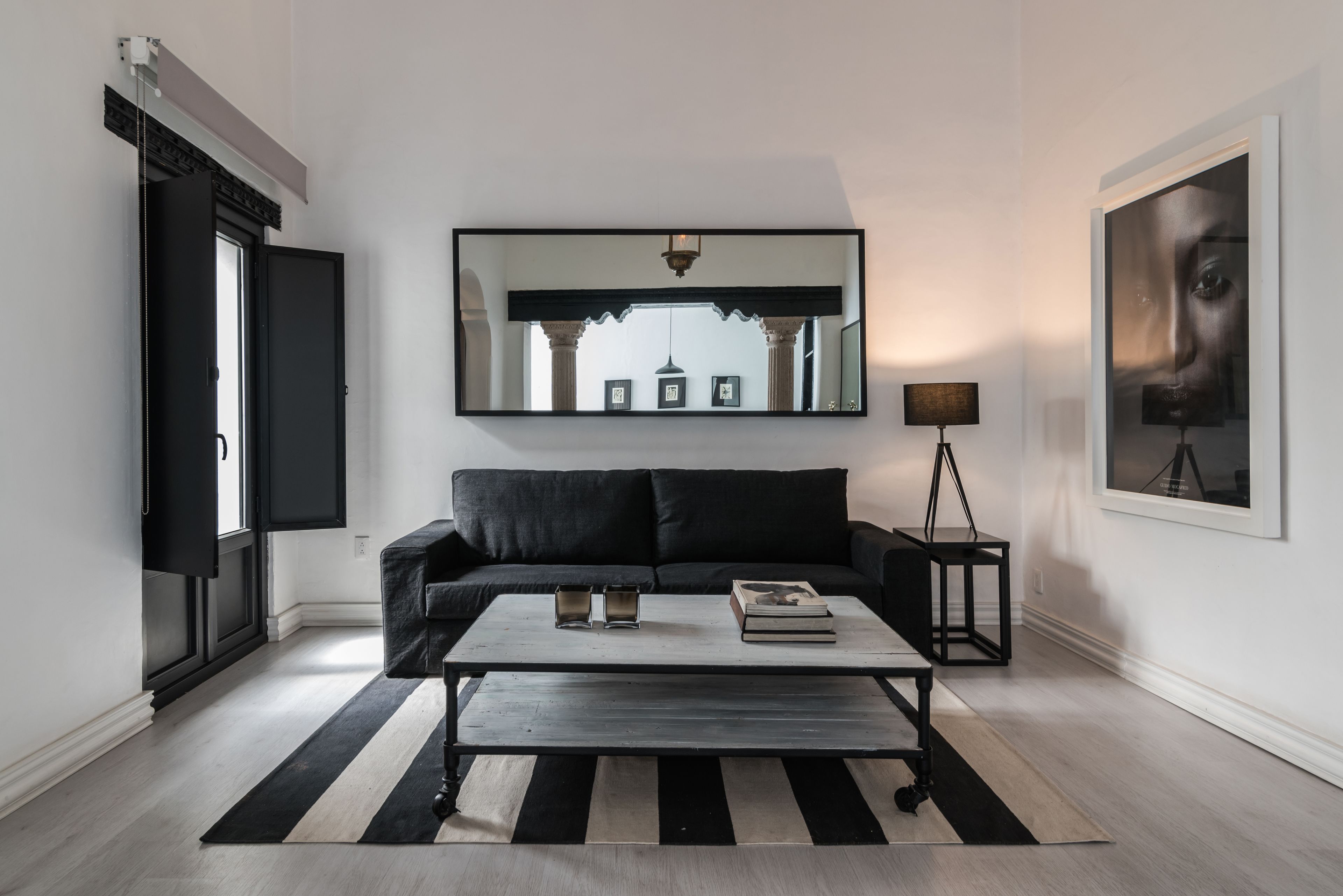 Ein modernes Wohnzimmer mit einem schwarzen Sofa, einem Couchtisch, einem schwarz-weiß gestreiften Teppich, einer Stehlampe und einem großen Spiegel an der Wand.