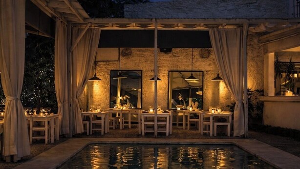 Un área de comedor al aire libre con mesas y sillas, una piscina y una sección cubierta con cortinas.