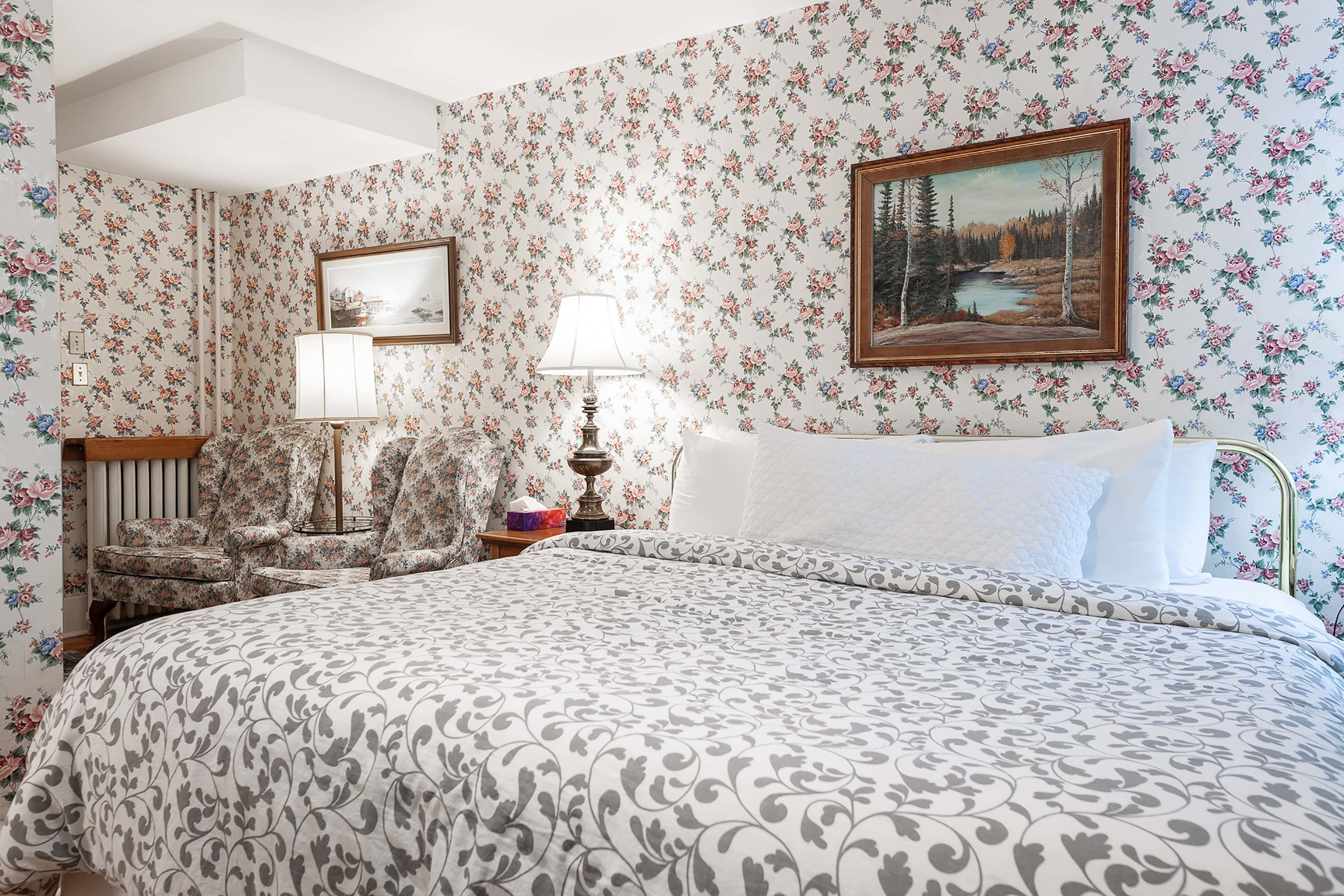 Un dormitorio con papel tapiz floral, una cama con colcha estampada, una mesita de noche con lámpara y un cuadro en la pared.