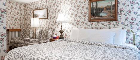 Un dormitorio con papel tapiz floral, una cama con colcha estampada, una mesita de noche con lámpara y un cuadro en la pared.