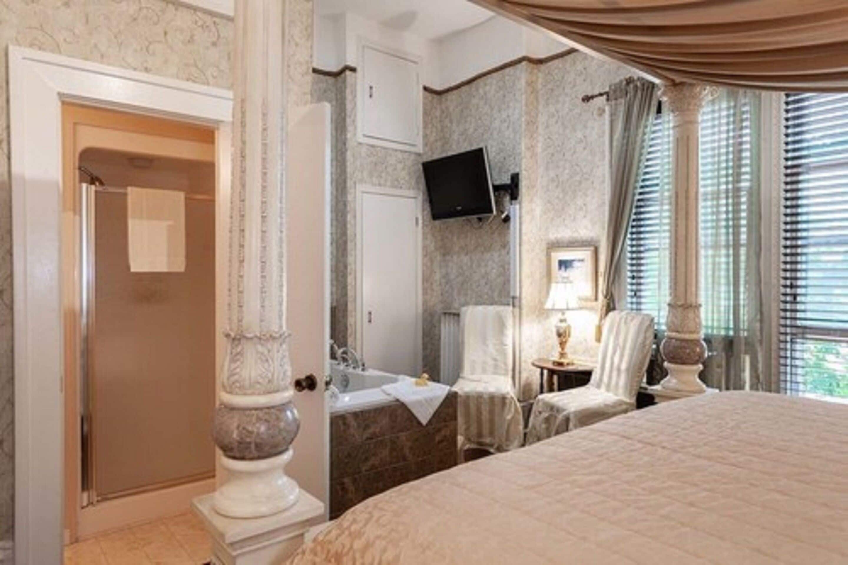A hotel room with a large bed, a bathroom with a bathtub, a TV mounted on the wall, and a window with blinds.