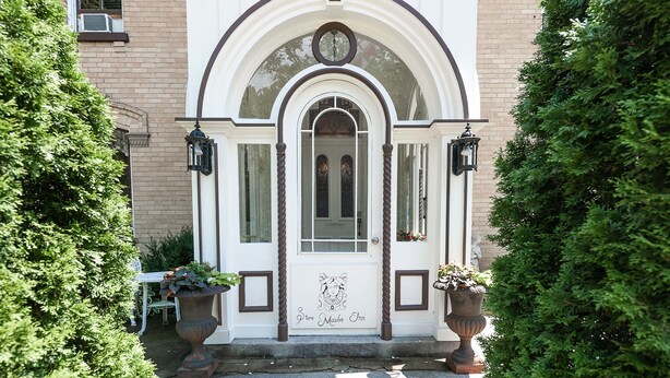 A white building with a central arched entrance, two potted plants on either side, and a small table with chairs outside.