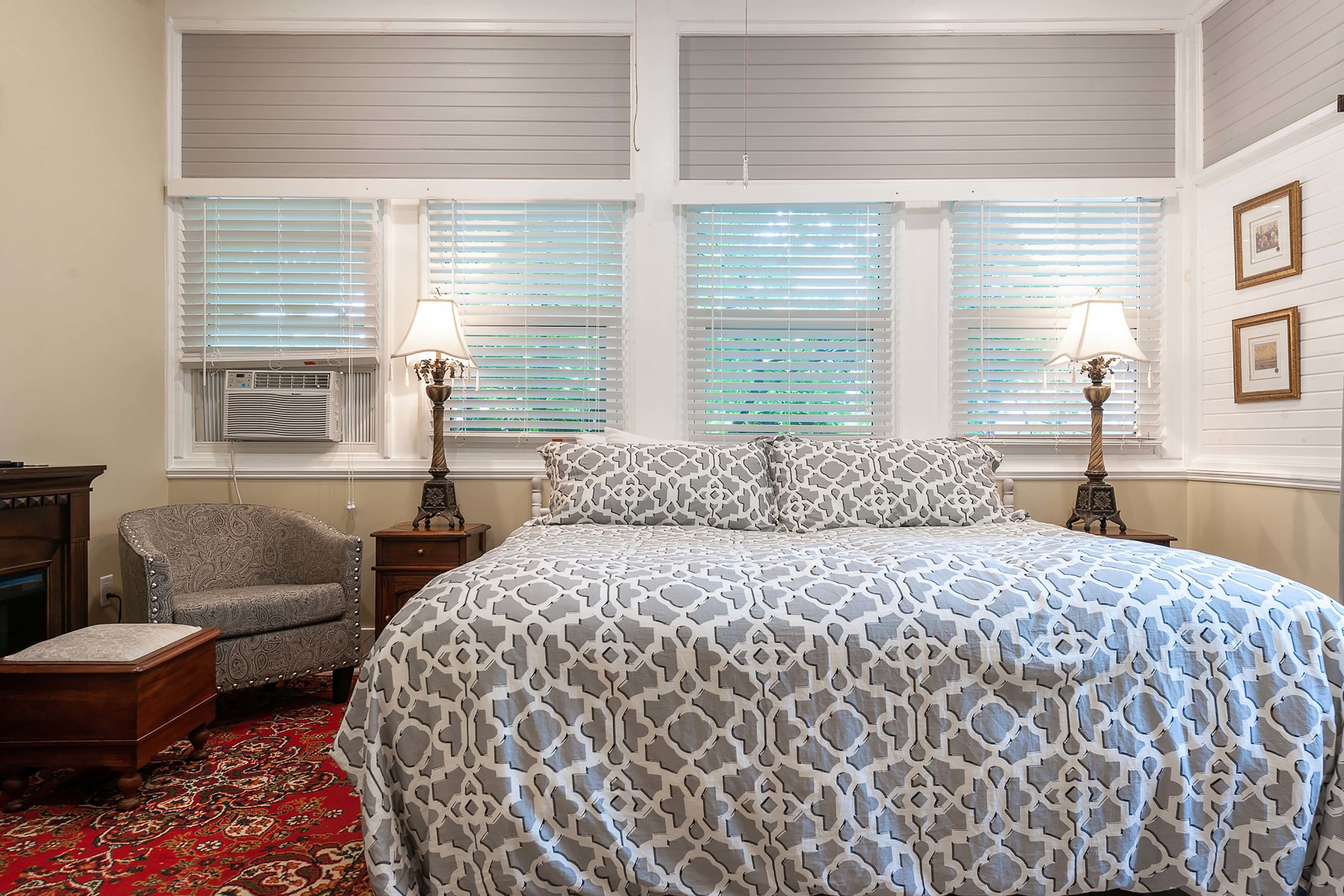 A bedroom with a patterned bedspread, two bedside lamps, a chair, a small table, and a window with blinds.