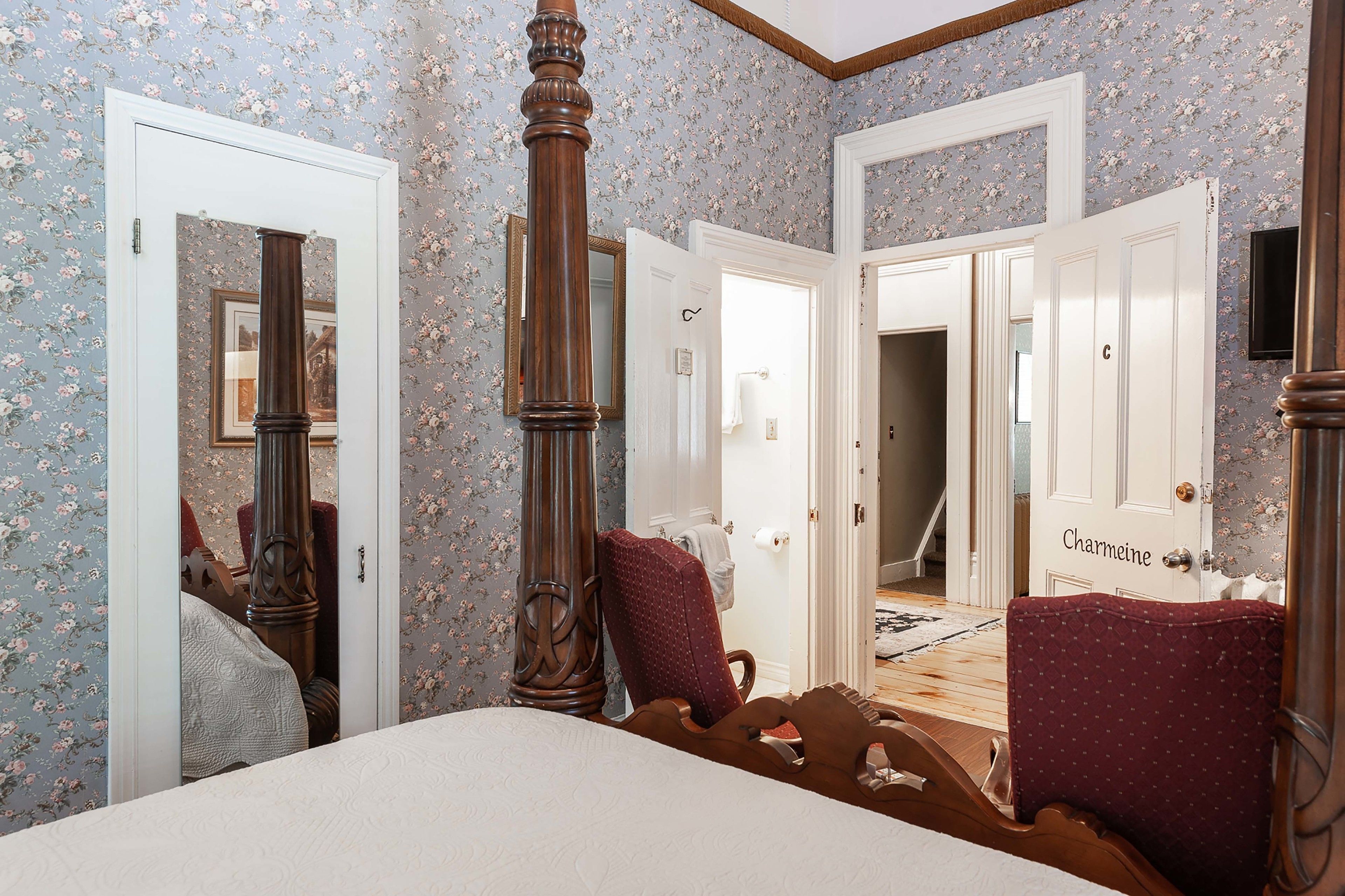 Un dormitorio con una cama grande, una silla de madera y un armario. Hay dos puertas que dan a otras habitaciones, una de ellas con la etiqueta "Charmeine".
