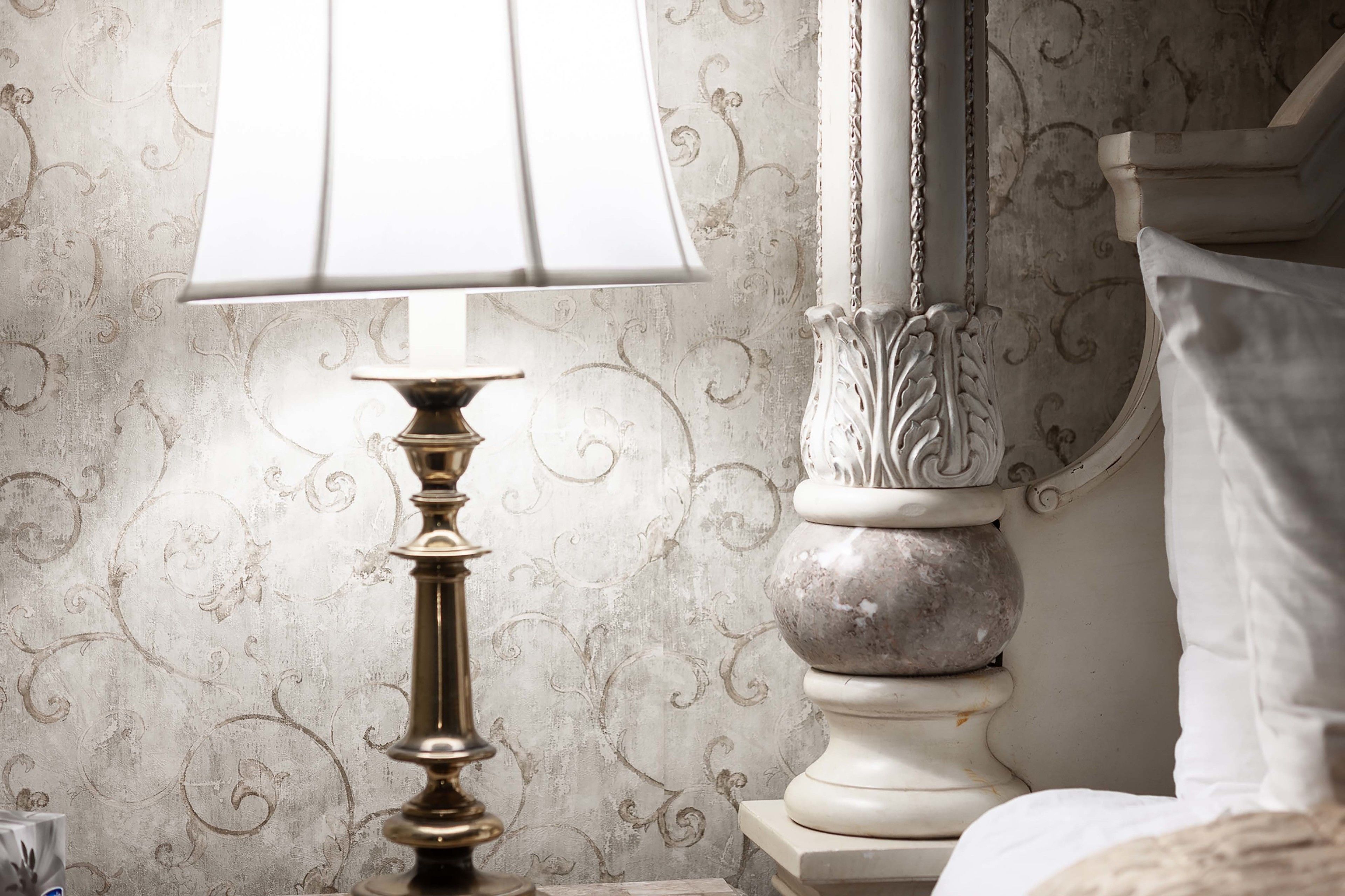 A lamp with a white shade on a bedside table, a decorative wall with a floral pattern, and a white bed with a pillow.