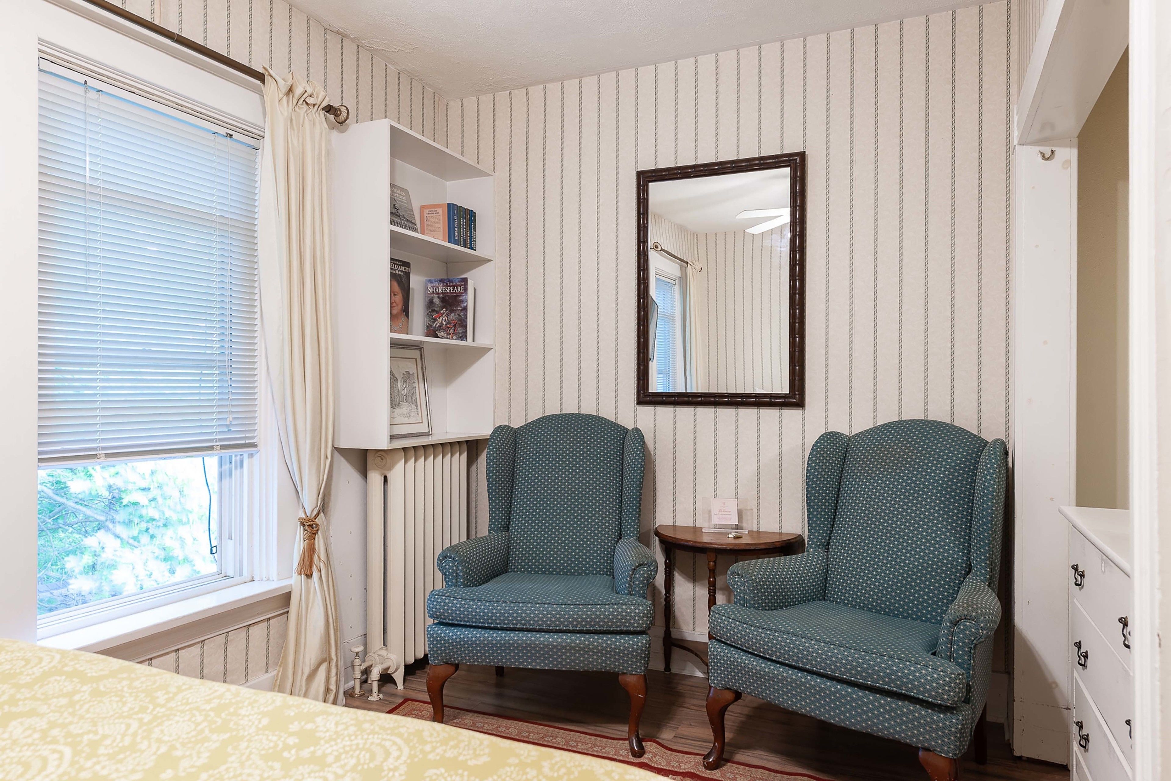 A room with striped wallpaper, two patterned armchairs, a small table, a bookshelf, and a window with curtains.