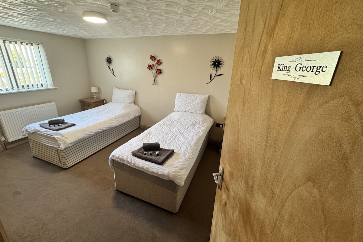 Et hotelværelse med to senge, en radiator, et vindue med rullegardiner og et navneskilt på døren.