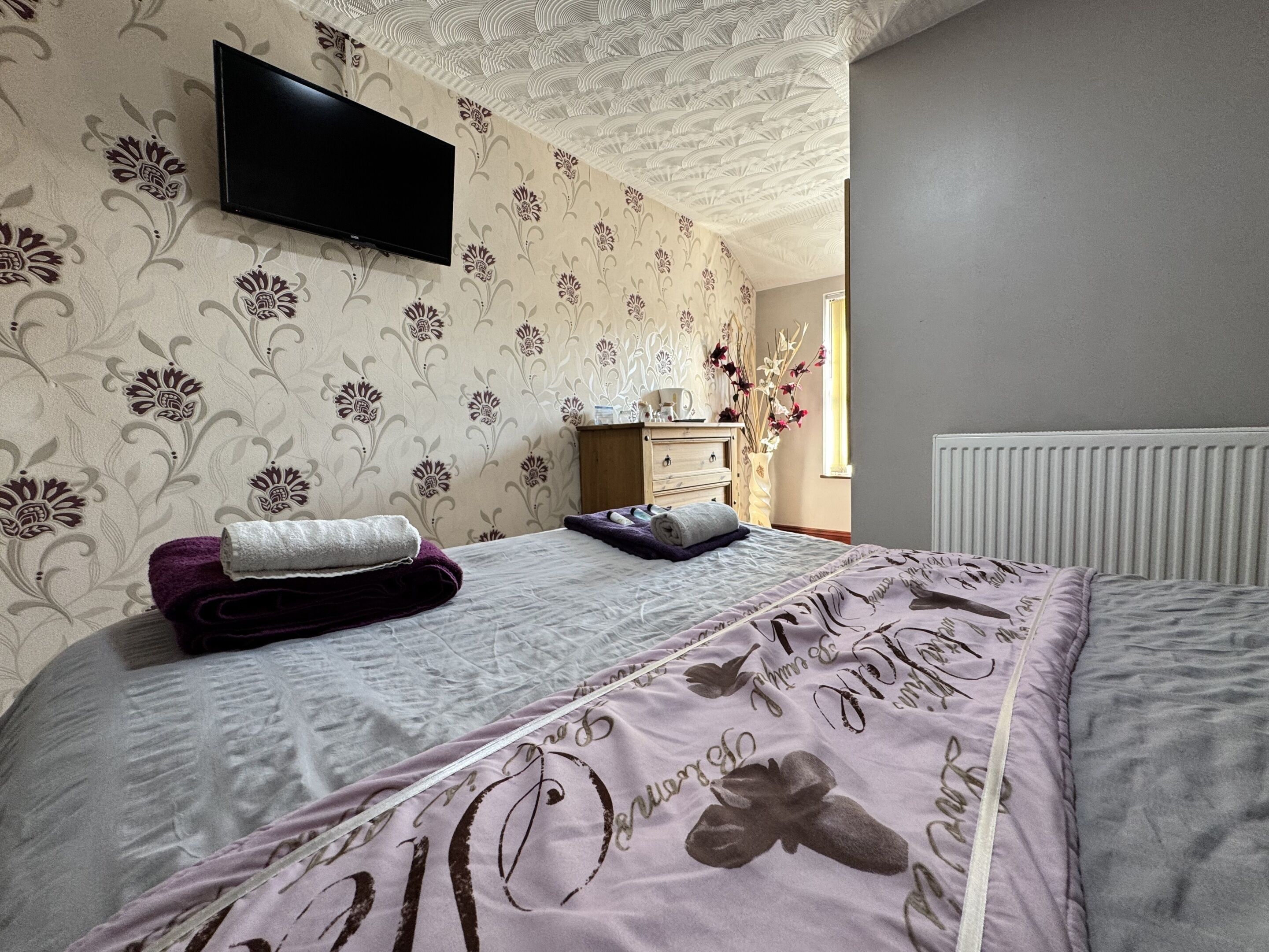 Una cama bien hecha con una colcha estampada con motivos florales y una toalla enrollada encima. Un televisor de pantalla plana está montado en la pared sobre la cama.