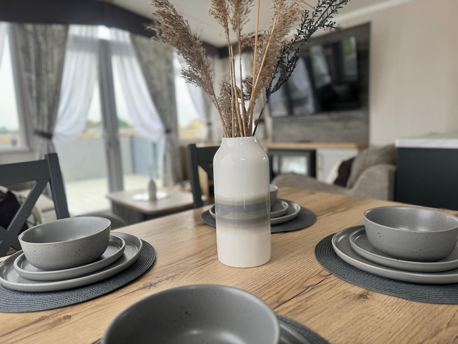A dining table with grey plates and bowls, a vase with dried plants, and a modern kitchen in the background.