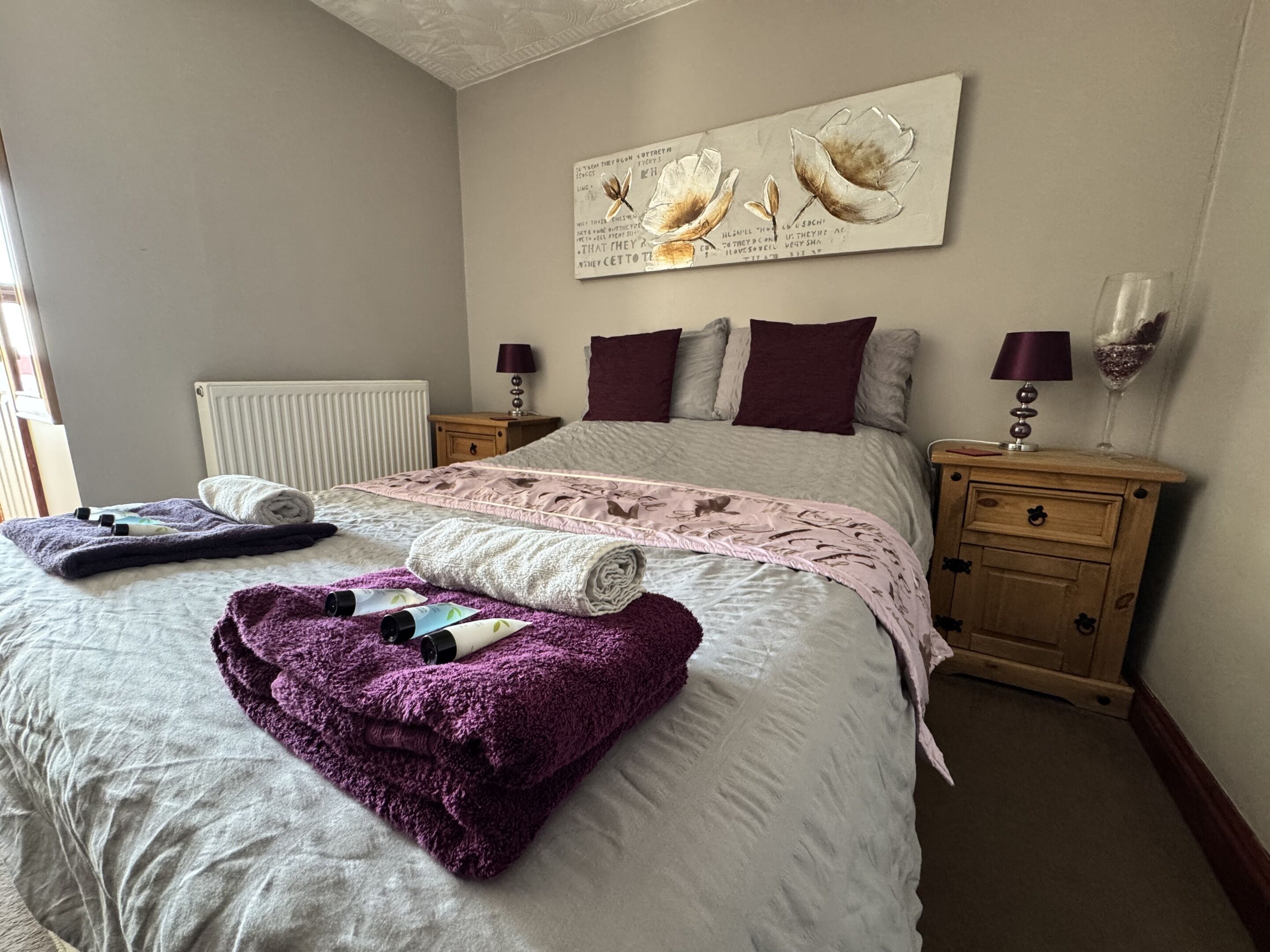 A neatly made bed with a purple blanket and rolled towels, a bedside table with a lamp, and a wall with a floral artwork.