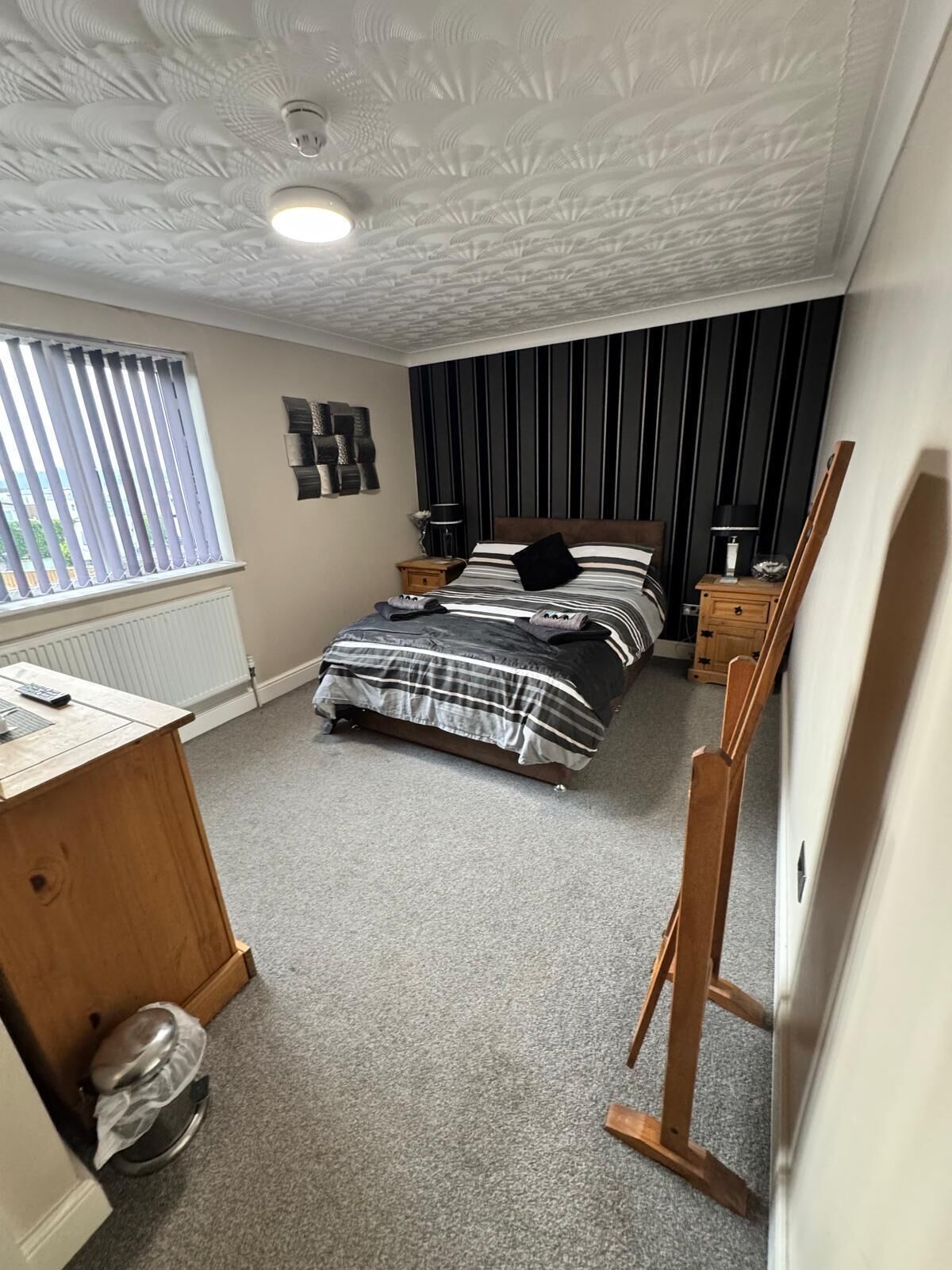 A bedroom with a bed, a desk, a radiator, a window with blinds, and a ceiling with a textured design.