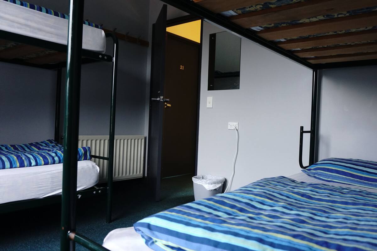 Een slaapzaal met stapelbedden, een radiator, een deur met een nummer en een spiegel aan de muur.