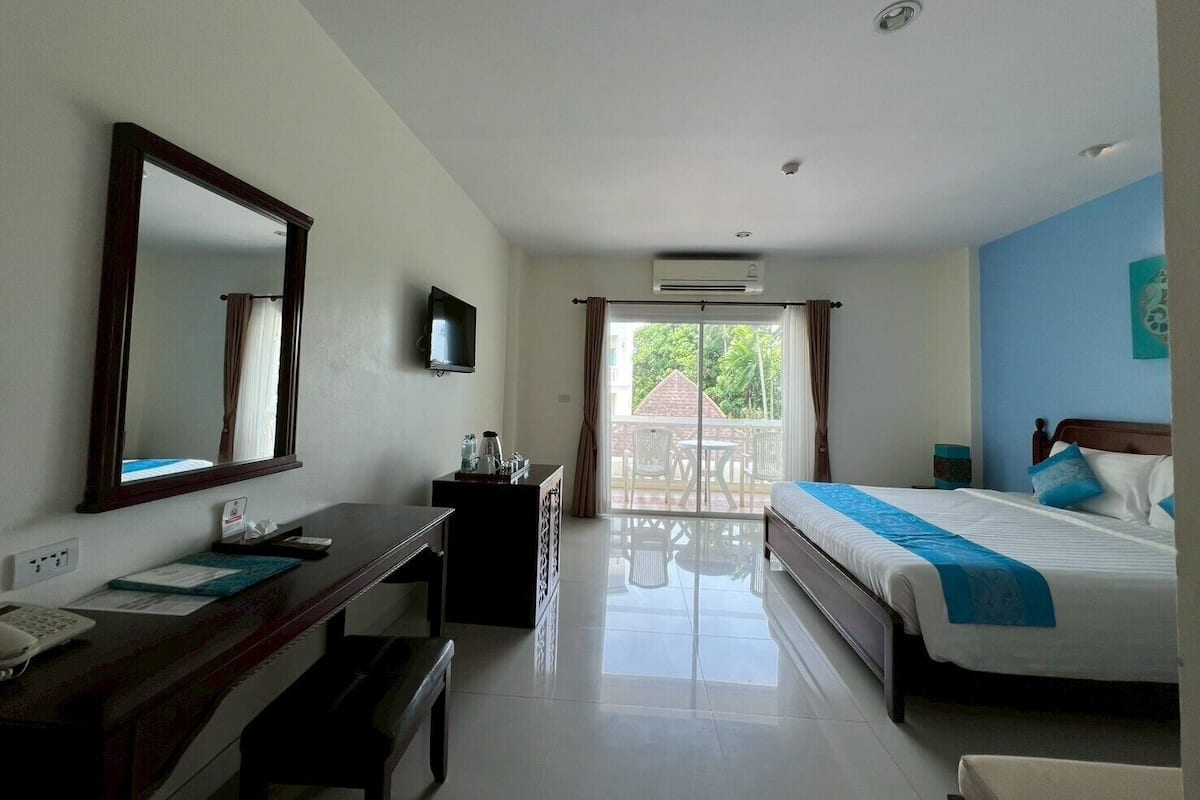 A hotel room with a bed, a desk, a mirror, a TV, and a balcony with a view of greenery.