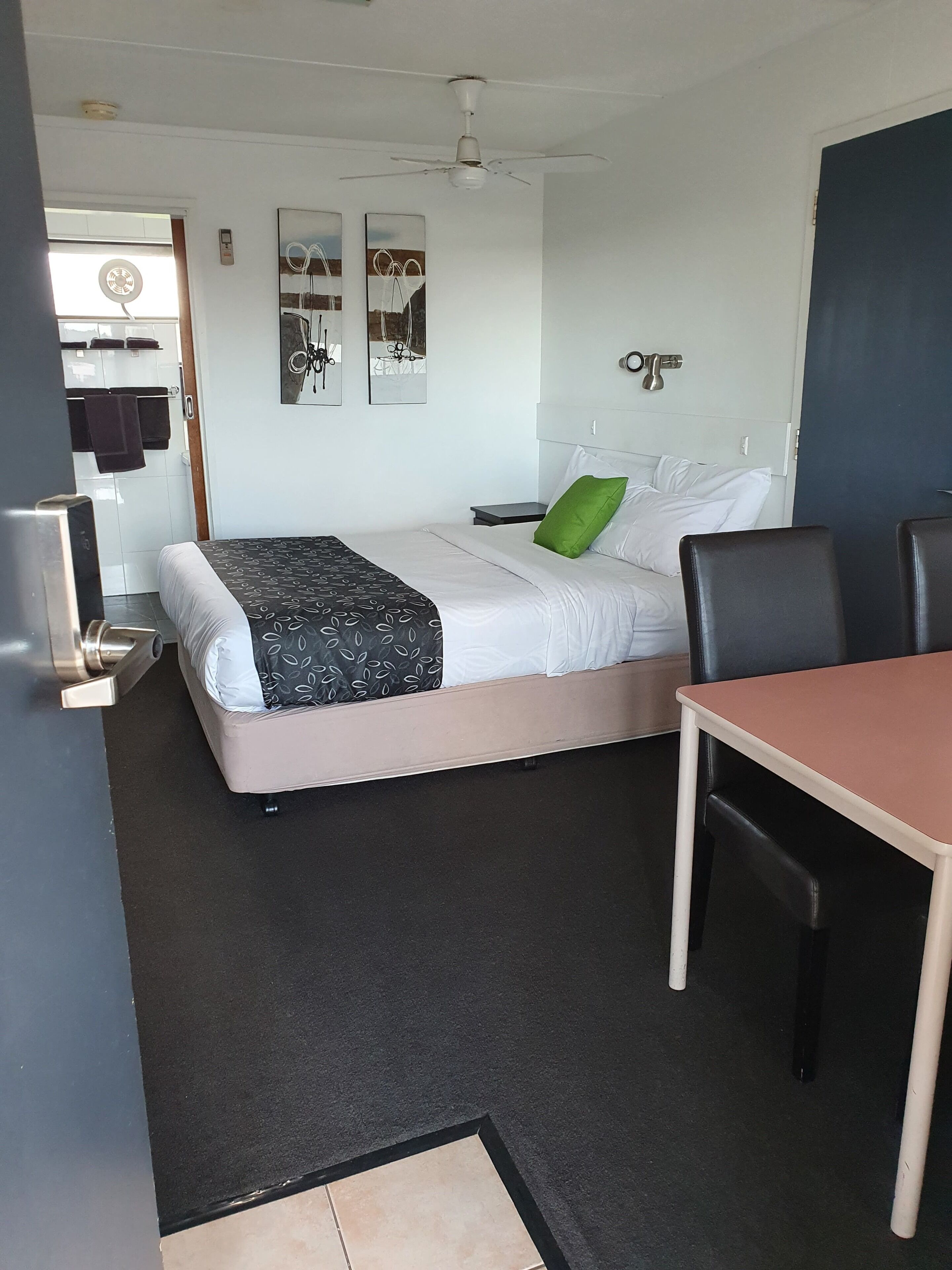 A hotel room with a bed, a desk, a chair, a small table, a ceiling fan, and a wall-mounted light.