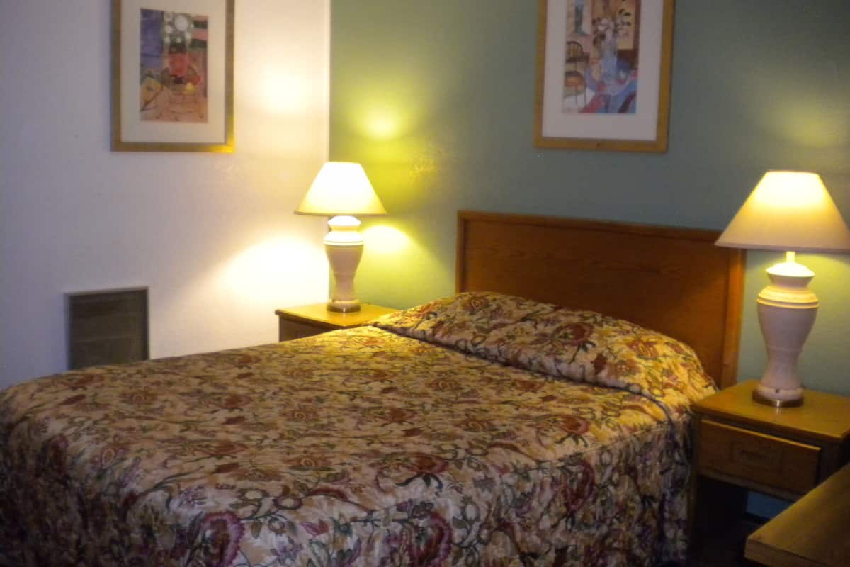 Cama bem arrumada com roupa de cama floral, duas luminárias de cabeceira e quadros nas paredes.