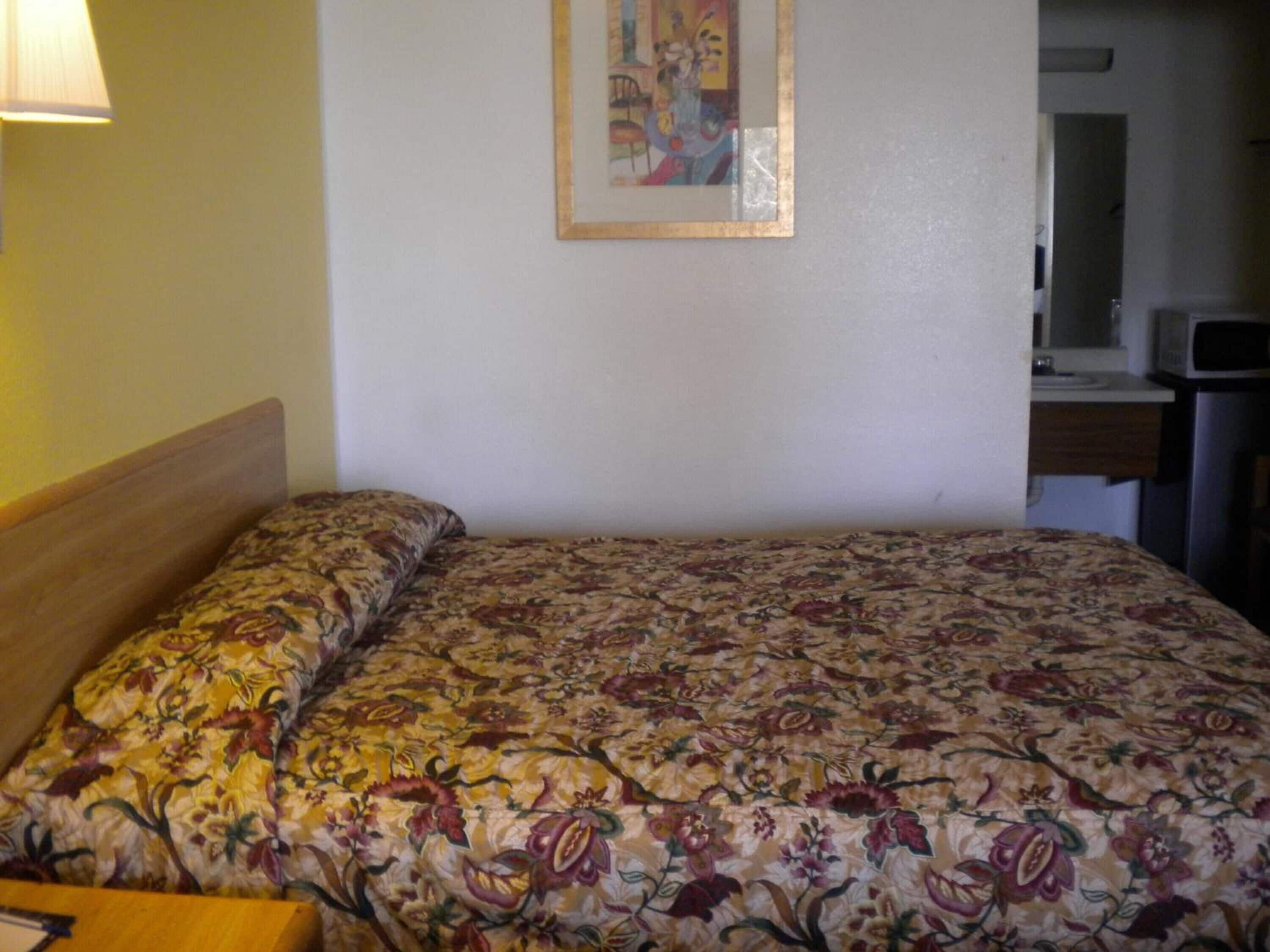 A hotel room with a bed, a nightstand, a lamp, a painting on the wall, and a kitchenette area.