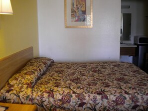 A hotel room with a bed, a nightstand, a lamp, a painting on the wall, and a kitchenette area.