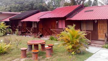 Une rangée de cabanes en bois aux toits rouges, chacune dotée d’un porche et d’une porte. On y trouve des tables et des chaises d’extérieur, ainsi que diverses plantes qui les entourent.