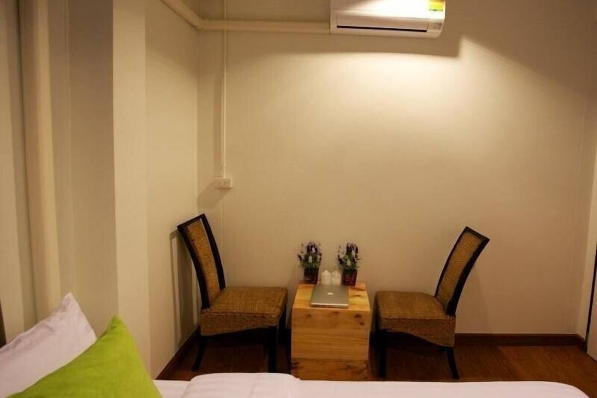 Una habitación pequeña y bien iluminada con una cama, dos sillas, una mesita con una computadora portátil y un equipo de aire acondicionado en la pared.