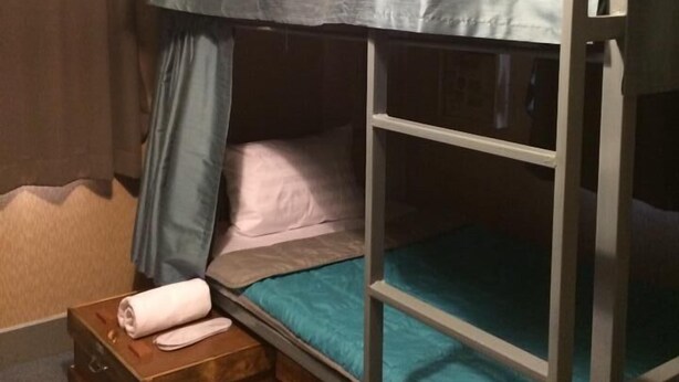 A bunk bed with a metal frame, a blue mattress, and a grey canopy. There is a wooden chest of drawers with a rolled-up towel on top. - Nap Box Hostel (Chiang Mai)