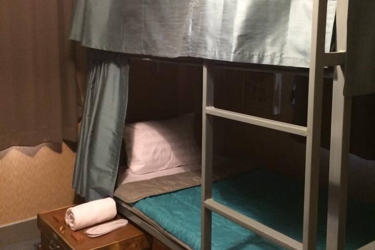 Una cama litera con estructura metálica, un colchón azul y un dosel gris. Se observa un aparador de madera con una toalla enrollada encima.