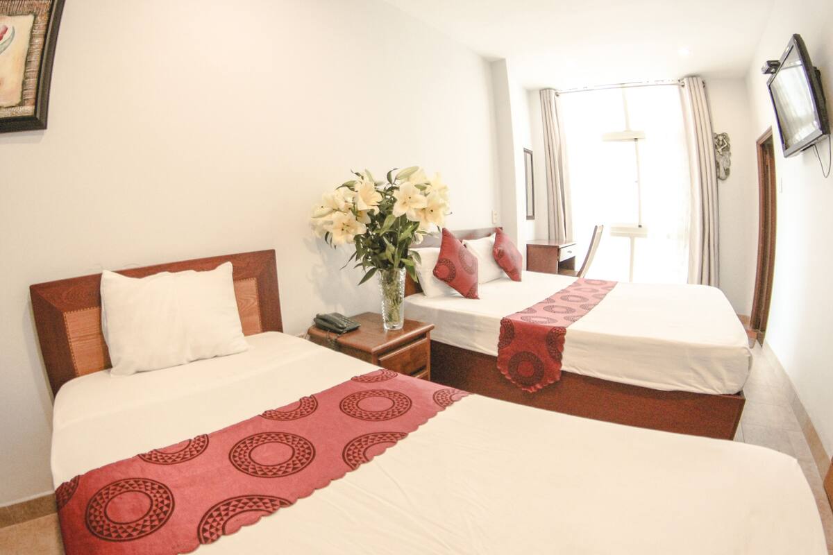 Ein Hotelzimmer mit zwei Betten, einem Nachttisch mit Blumen in einer Vase, einem Fenster mit Vorhängen und einem an der Wand befestigten Fernseher.