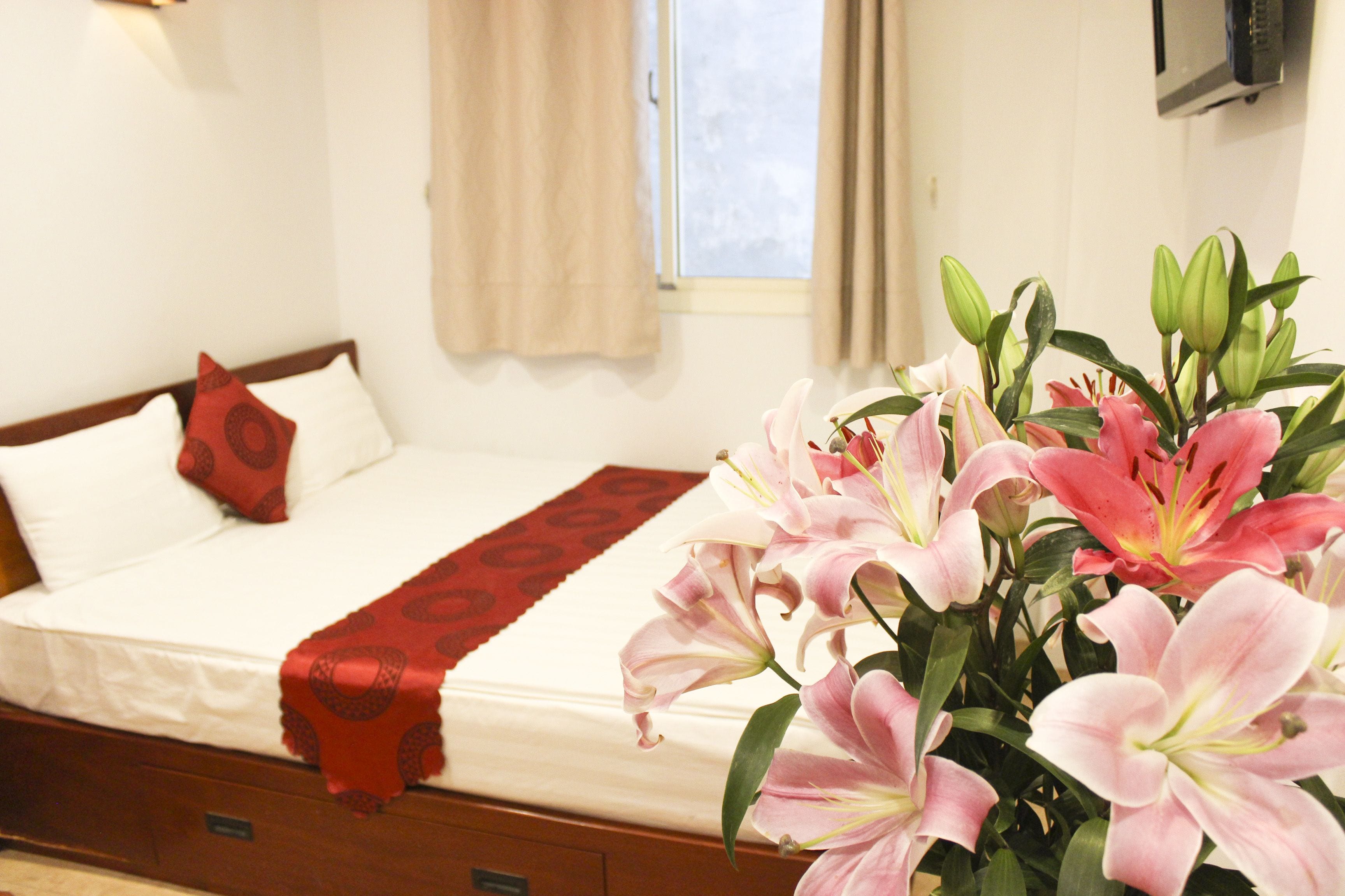 Une chambre d’hôtel avec un lit, un plaid rouge et un vase rempli de lys roses.