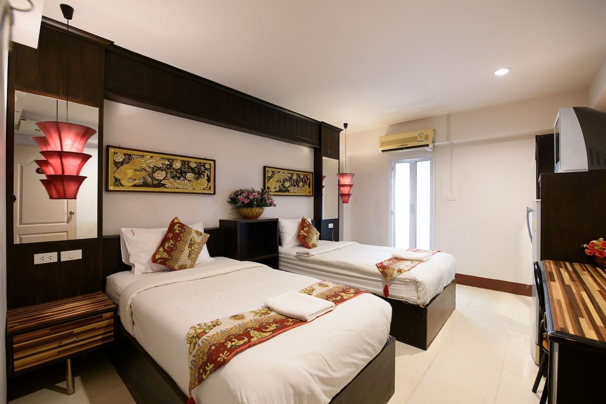 Habitación de hotel con dos camas, una mesita de noche de madera, un escritorio, un espejo, detalles decorativos en la pared y una ventana con unidad de aire acondicionado.
