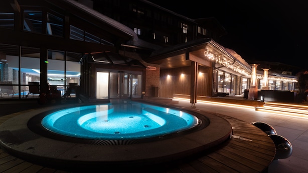 Een buitenzwembad met verwarming, een houten terras, een gebouw met grote ramen en een bar met verlichting.