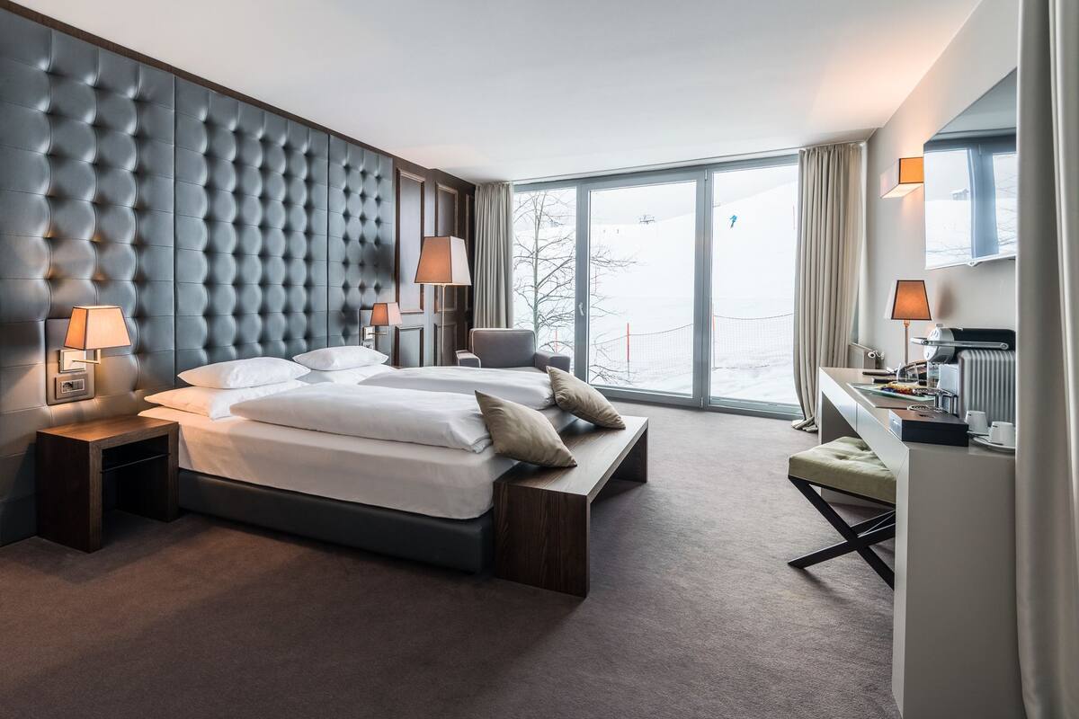 Habitación de hotel moderna con una cama grande, dos mesitas de noche, un escritorio con una silla y un amplio ventanal con vistas al exterior.
