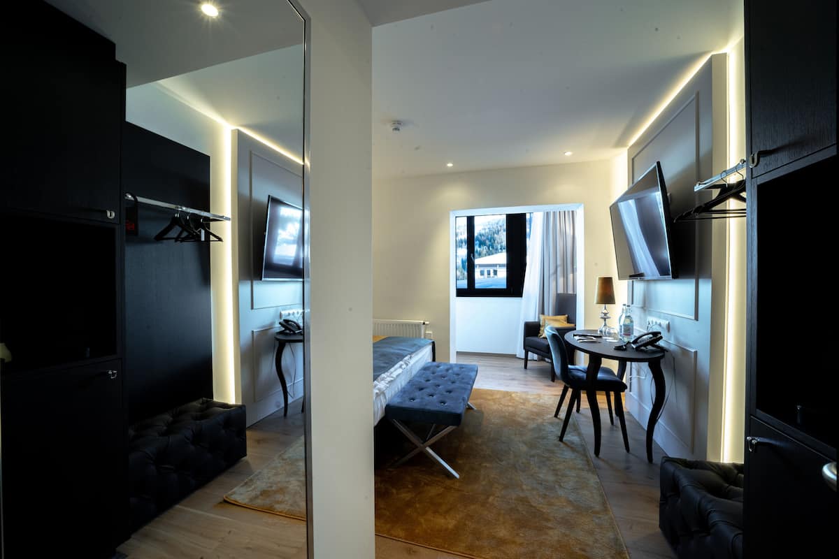 Una habitación de hotel moderna con una cama, una zona de estar con una mesa y sillas, un televisor montado en la pared y un ventanal con vistas al exterior.