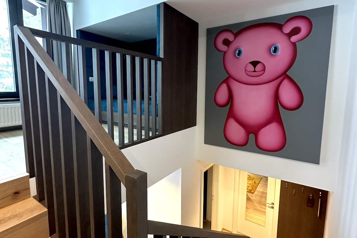 Una escalera moderna con escalones de madera y barandillas oscuras, que conduce a un rellano con un cuadro grande de un oso rosa.