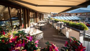Een overdekt terras met tafels en stoelen, grote ramen en uitzicht op de straat.