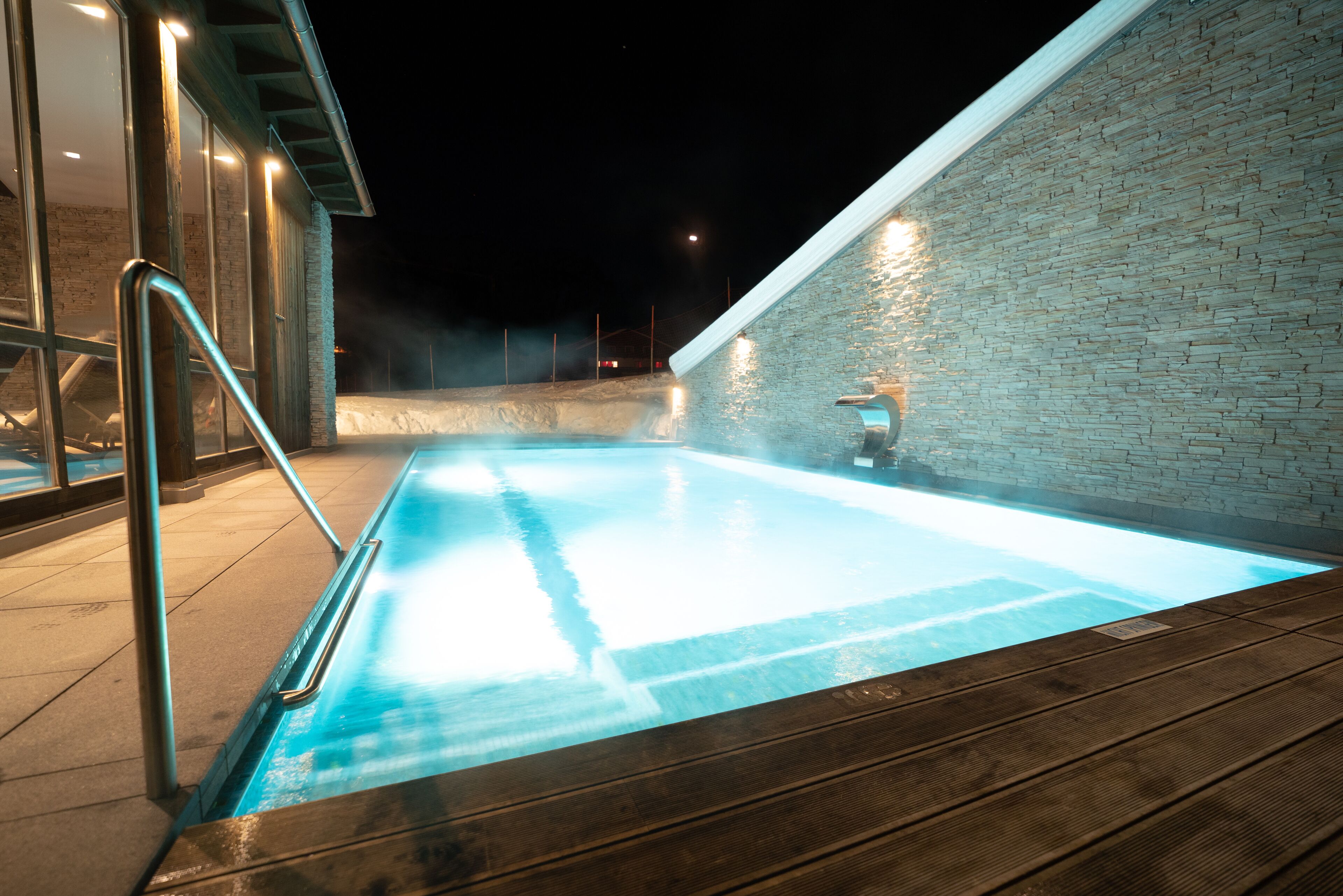 Une piscine intérieure à l’eau claire, entourée d’une terrasse en bois et d’un mur en briques.
