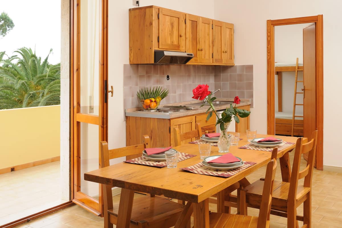 A dining area with a wooden table set for four, a kitchen with wooden cabinets, and a balcony with plants.