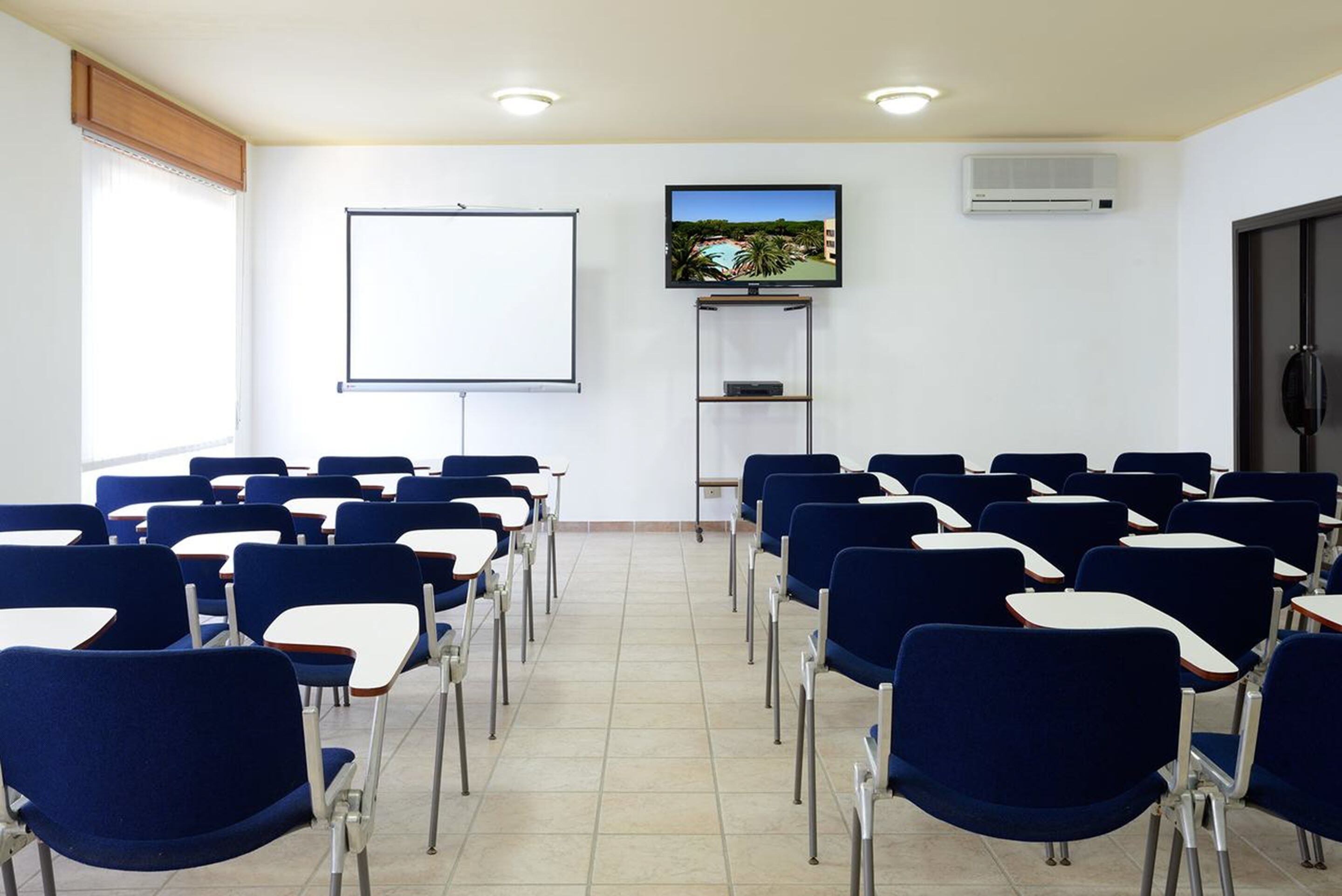 A classroom with rows of blue chairs and white desks, a whiteboard, a television on a stand, and an air conditioning unit on the wall.